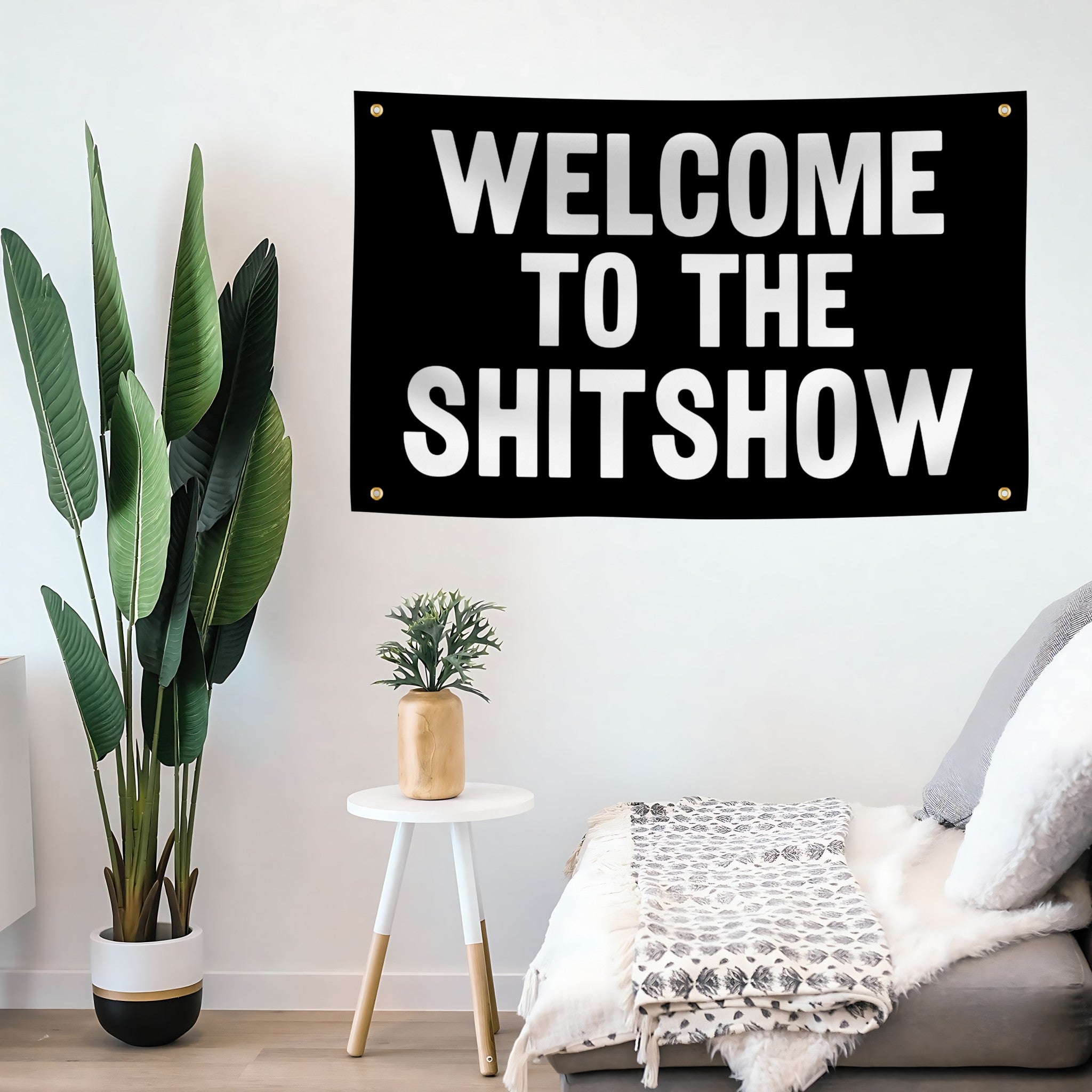 A black rectangular wall banner with bold white text reading 'WELCOME TO THE SHITSHOW' hanging on a white wall above a bed, with a potted plant and small side table nearby.